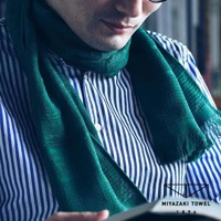 一位男士頸部圍繞著一條深綠色、帶有流蘇的今治匹馬棉圍巾，圍巾材質輕薄，露出內搭的藍白條紋襯衫。