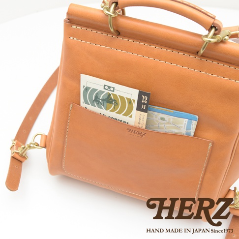 職人手作皮革屋瓦風後背包，淺棕色皮革，包身縫線明顯，正面有一個較大的置物袋，內有雜誌和地圖。