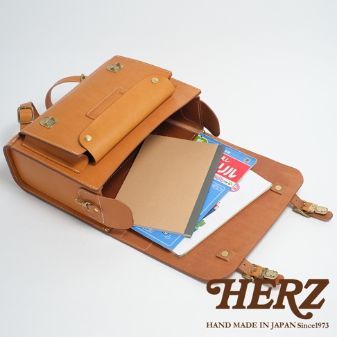 一個棕色皮革製成的橫式書包，包蓋打開，裡面放著幾本筆記本和書籍，包包的右下角有HERZ的商標。