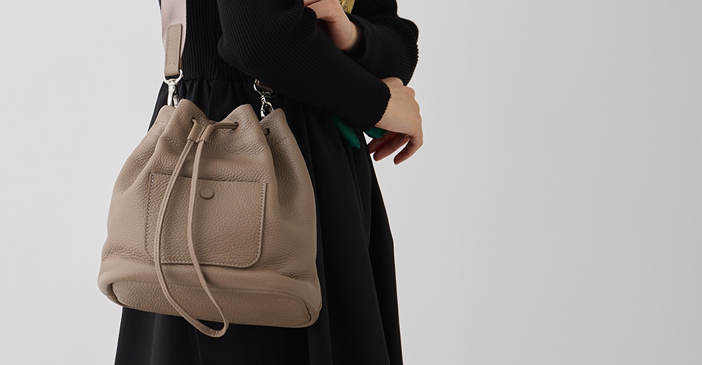 一個戴著黑色上衣和黑色連衣裙的女人，左肩上掛著一個米色皮革束口水桶包。