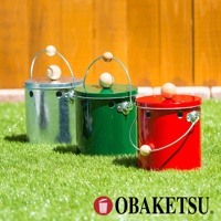 OBAKETSU 品牌標誌旁擺放著三個不同顏色的手工製蚊香桶，分別為金屬原色、綠色和紅色，放置於綠色草地上，背景為木質柵欄。