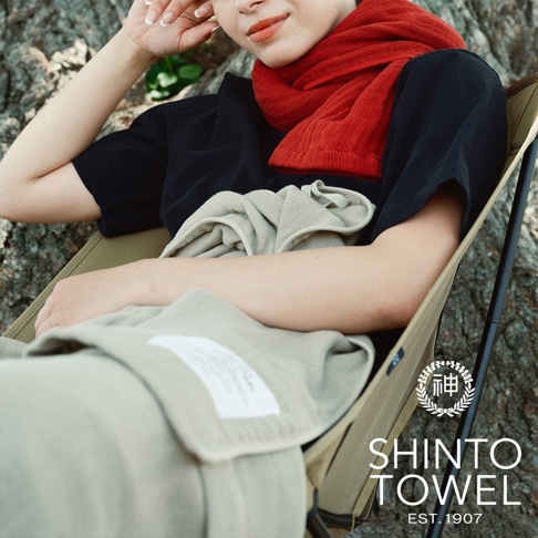 一名女子坐在一張卡其色露營椅上，身上蓋著一條淺卡其色的暖毯，頸部圍著一條紅色圍巾。暖毯質地柔軟，表面有細緻的紋理。