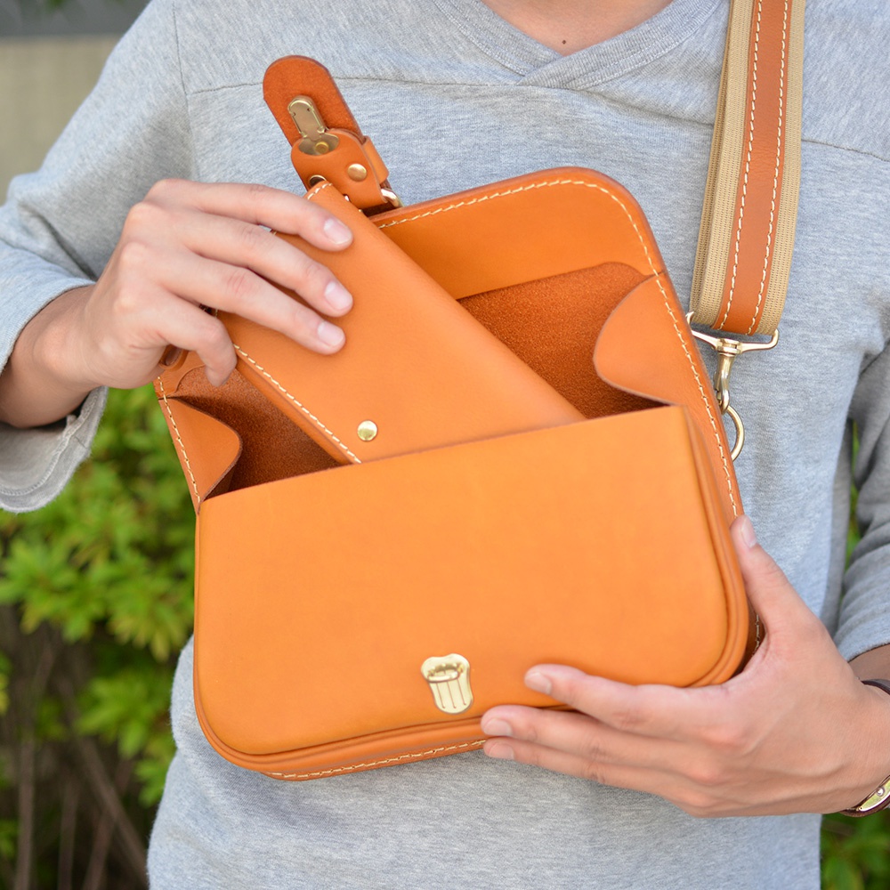 職人手作皮革單扣經典斜背包，展現溫潤的橘棕色皮革質感，搭配米色織帶與同色系縫線。包款中央配有金色單扣，內部可見置入的長夾。