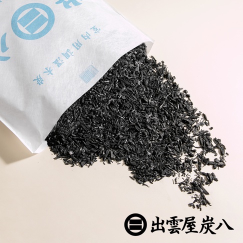 白色紙袋傾斜，倒出大量黑色碎狀木炭，木炭散落在淺米色背景上。紙袋上有藍色印刷文字與標誌。右下角有黑色圓形標誌和「出雲屋炭八」文字。