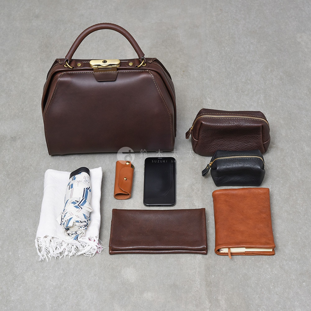 職人手作深棕色皮革兩用口金手提包，附有各式皮革配件：一個咖啡色小化妝包、一個黑色小零錢包、一個棕色鑰匙包、一個黑色手機、一個深棕色長夾，以及一個淺棕色筆記本。