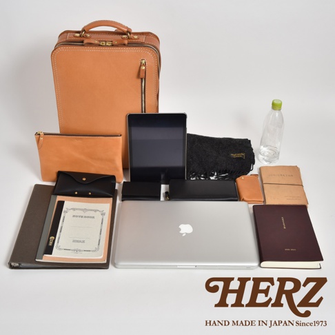 一個職人手作的淺棕色皮革電腦後背包，擺放著一個相同材質的化妝包、一個黑色皮夾、一個黑色筆記本、一個平板電腦、一個銀色筆記型電腦，以及一條黑色圍巾、一個淺棕色皮製卡夾、一本棕色筆記本和一瓶水。