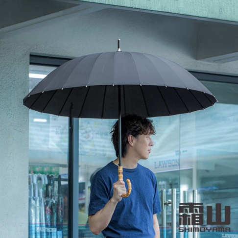 一名男子站在便利商店門口，手中握著一把日本霜山品牌出品的黑色晴雨兩用傘，傘面設計為堅固的二十四骨架結構，搭配具備質感的天然竹節彎曲傘柄，背景可見便利商店玻璃門與內部陳列的雨傘架。