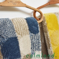 兩款北歐風地墊，一款為藍色與米色拼接格紋，另一款為黃色與米色拼接格紋，皆為柔軟的絨毛材質，由木製衣架懸掛展示。