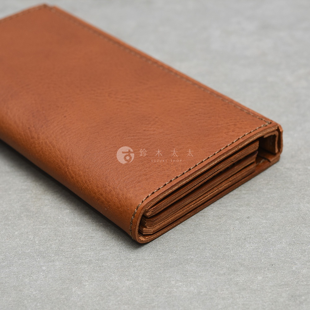 這是一個日本HERZ職人手作的棕色皮革長夾，採用Organ系列設計，擁有厚實且質感細膩的皮革紋理，側面展示出多層次的摺疊收納結構與精緻的縫線細節，主體表面印有鈴木太太的品牌標誌。