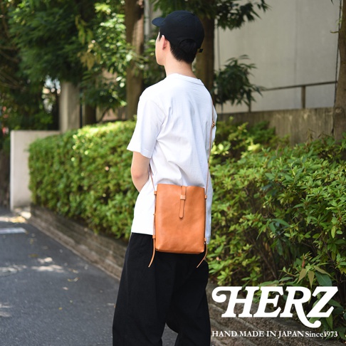 一位背對鏡頭的男性，肩背一個由日本品牌 HERZ 手工製作的淺棕色直式皮革側背包。該包款設計簡約輕薄，採用柔軟皮革材質，搭配獨特的皮帶釦設計，適合隨身攜帶長夾與手機等日常用品。背景為充滿綠意的戶外街道。