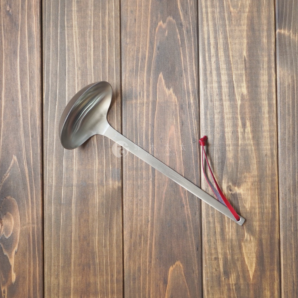 一個銀色不鏽鋼萬用湯勺，勺身呈橢圓形，帶有細長的勺柄，勺柄末端繫著一小撮紅色細繩。