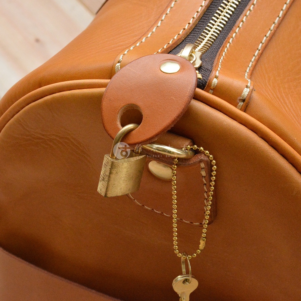 焦糖色皮革波士頓包，配有金色拉鍊、鎖頭與鑰匙。皮革表面有細緻縫線，並附有皮革製的鎖頭掛牌。