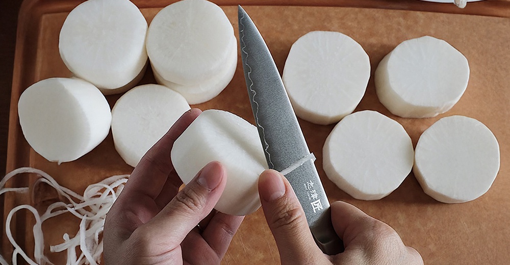 一把銀色金屬材質的Chef's knife主廚刀，刀刃呈波浪狀，正在切削一塊白色的白蘿蔔。周圍散落著幾片已切好的白蘿蔔圓片，以及一些刨成細絲的白蘿蔔。