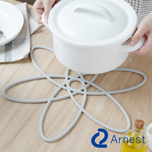 一個淺灰色、花瓣狀的矽膠鍋具防刮保護墊，上面放置著一個白色帶蓋的鍋具，背景為木質桌面。