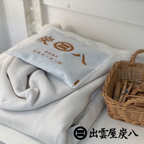 一疊米白色的厚織物折疊放置在桌面，上方放著一袋印有出雲屋炭八品牌標誌與文字的白色方形除濕木炭包，右側有一個放置木夾的編織籃，右下角則標示著出雲屋炭八的品牌名稱與標誌。