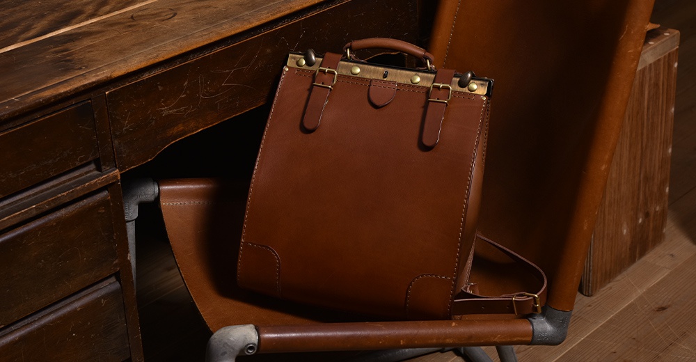 一個復古風格的棕色皮革後背包，配有金色金屬口金、搭扣和提把，放置在木製椅子上，背景為木製桌子。