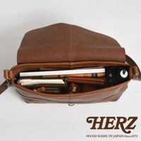 日本HERZ職人手作皮革細扣帶馬鞍包，展示打開的包內部裝有書籍、錢包、記事本及雨傘等物品。