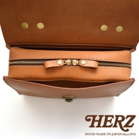 日本HERZ職人手作皮革多功能收納單肩包，近距離拍攝，展示了包包的拉鍊和翻蓋細節，包身為焦糖棕色，品牌標誌HERZ及產地字樣位於畫面下方