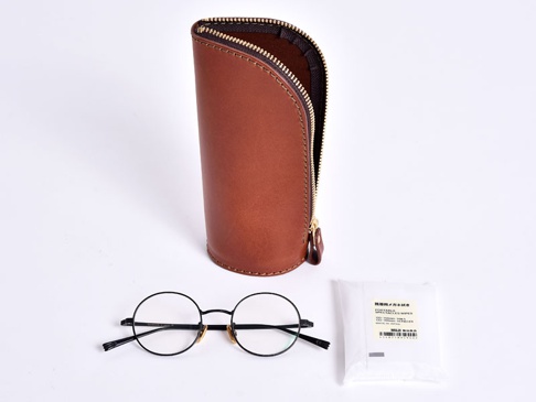 職人手作皮革直立式收納筆袋，經典棕色，附有拉鍊開合。下方擺放一副黑色圓框眼鏡及一包眼鏡清潔濕巾。