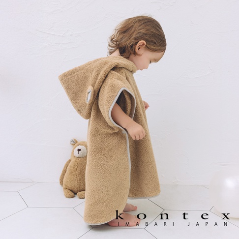 穿著淺棕色毛絨材質、帶有熊耳造型連帽的斗篷式浴巾，浴巾邊緣有灰色滾邊。