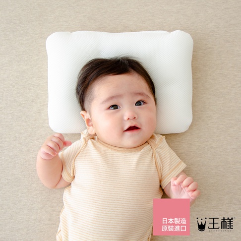 一名約 0-6 個月大的嬰兒，頭部躺在一個白色網狀材質的嬰兒枕上，嬰兒穿著一件淺黃色條紋連體衣，背景為米色布料。