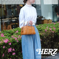 一位女性手提日本HERZ品牌駝色皮革迷你波士頓包，穿著白色寬袖上衣與牛仔寬褲，背景是綠色植物與店家櫥窗