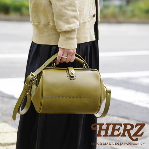 一只橄欖綠皮革的職人手作口金醫生包，附有可調節的單肩背帶。包身線條簡潔，配有金色旋轉鎖扣。