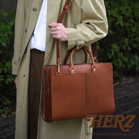 職人手作皮革雙口袋兩用商務公事包，採用鞣製皮革製成，呈現溫潤的棕色調。包款設有雙前袋、手提把及可拆卸肩背帶，整體線條簡潔，適合商務場合。