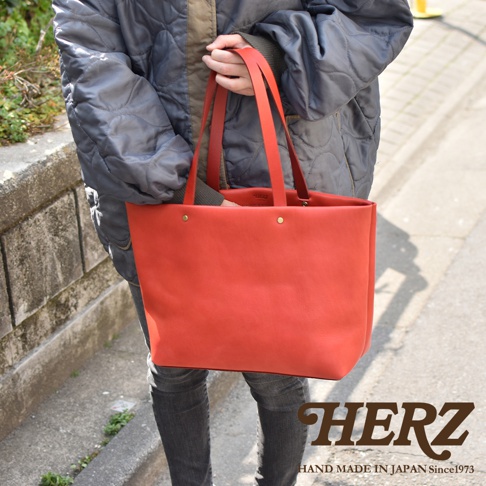 一位人士手持紅色皮革托特包，包包為簡約設計，有兩條皮革提把，包身正面有兩個鉚釘裝飾。