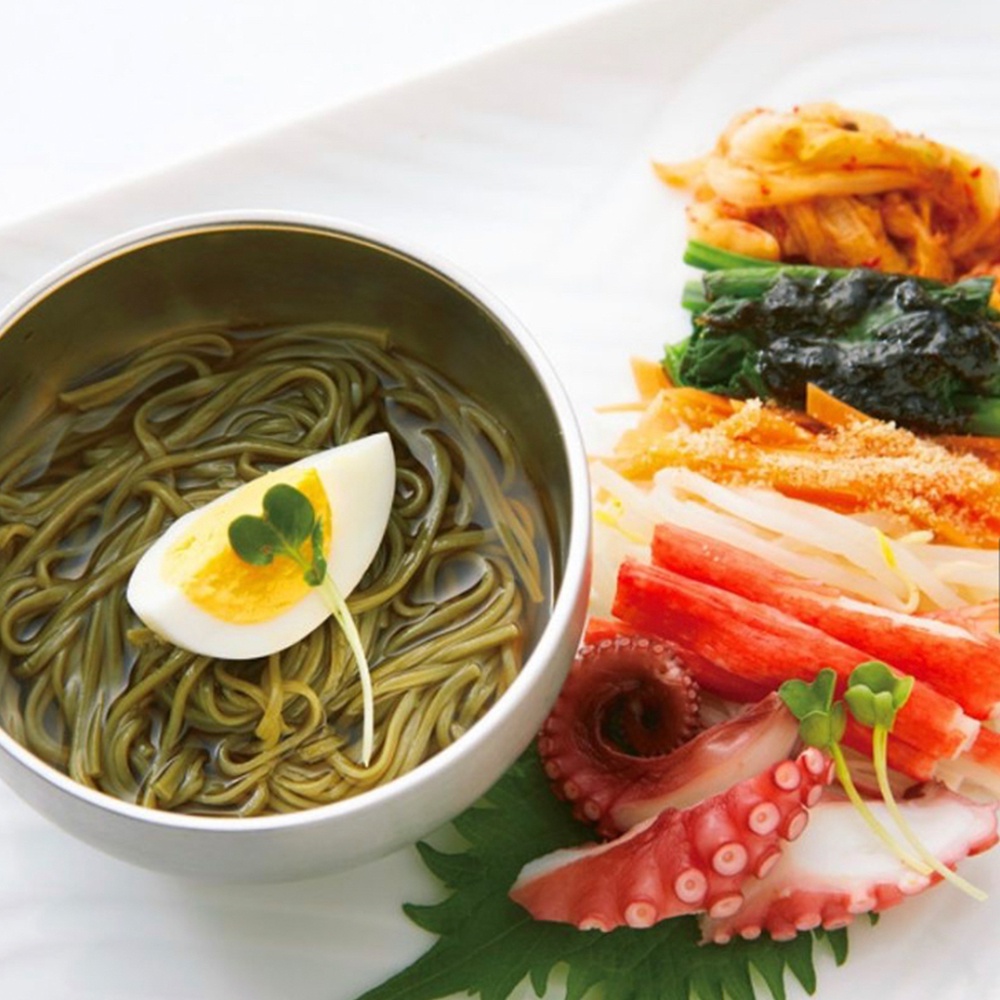 一碗裝有綠色蕎麥麵和半顆水煮蛋的銀色碗，旁邊擺放著章魚腳、蟹肉棒、豆芽菜、菠菜和泡菜。