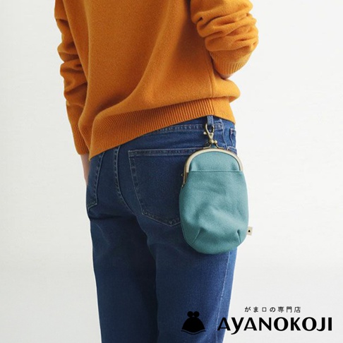 一個身穿橘色毛衣和藍色牛仔褲的人，側背著一個藍綠色的帆布腰掛口金包，包包掛在腰間的皮帶環上。