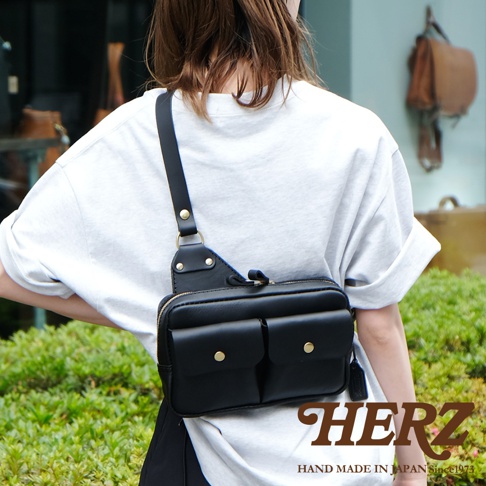 黑色皮革雙口袋斜背包，附有拉鍊主袋和兩個翻蓋前袋，配有金屬釦，可斜背或胸掛，展現職人手作質感。