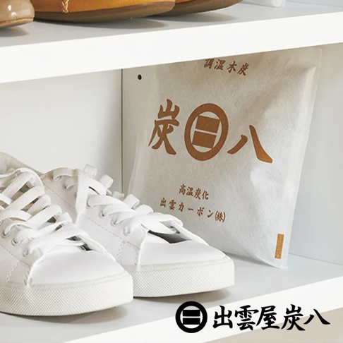 白色鞋櫃層架上放置一包印有出雲屋炭八標誌的方形調濕木炭，包裝採用白色不織布材質，旁邊擺放著一雙白色休閒運動鞋，整體呈現簡約清新的居家收納氛圍。