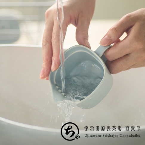 雙手在水槽邊清洗一個淡藍色的美濃燒一人用茶壺，水流正注入壺內，壺嘴處帶有濾網設計，畫面右下方印有宇治田原製茶場直賣部的品牌標誌。