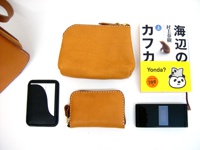 職人手作皮革翻蓋直式單肩包 (CP-96) 展示了兩個棕褐色皮革零錢包，一個黑色卡夾，一本黃色封面日文書籍，以及一部黑色摺疊手機。