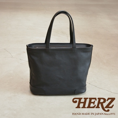 黑色皮革托特包，有拉鍊開口，兩個提把，底部有加固設計，包包下方印有HERZ字樣和HAND MADE IN JAPAN Since1973。