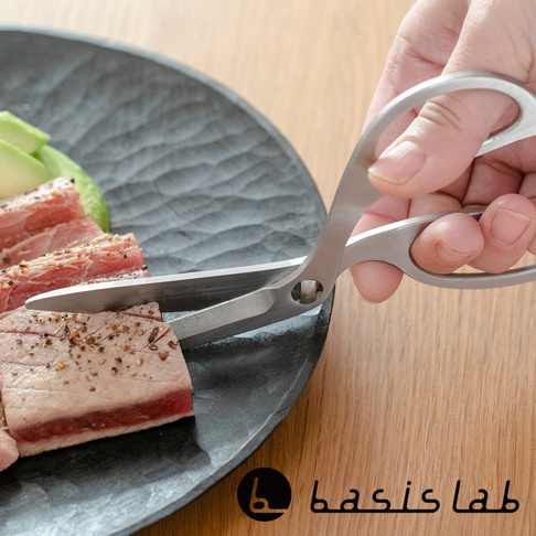 一隻手正使用一對銀色不鏽鋼料理剪刀，剪切盤中的肉塊，盤中還有切片的酪梨。剪刀具有可拆卸的設計，材質為光滑的拋光不鏽鋼。