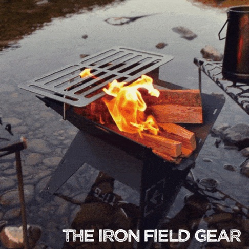 TAKI BEITA焚火台爐架套裝 S，由黑色金屬製成，內有燃燒的木柴和上方置放的銀色金屬烤網，整體放置於靠近水邊的戶外環境。