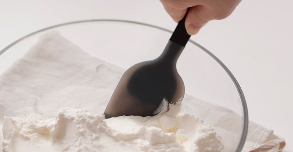 一支手正用黑色矽膠刮刀從裝有白色粉末的玻璃碗中舀取粉末，碗內鋪著白色布料。