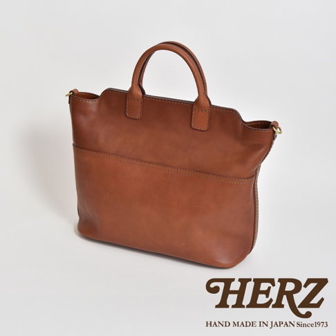 職人手作皮革大容量兩用托特包，採用溫潤的棕褐色皮革，包身線條簡潔，正面有橫向縫線裝飾，設有提把及側邊掛環，可肩背或手提。