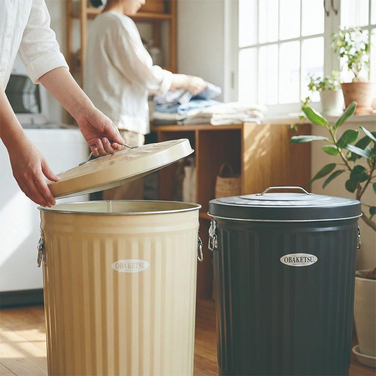 OBAKETSU 兩個附輪垃圾桶，一個米色，一個黑色，其中米色垃圾桶的蓋子被一隻手提起。