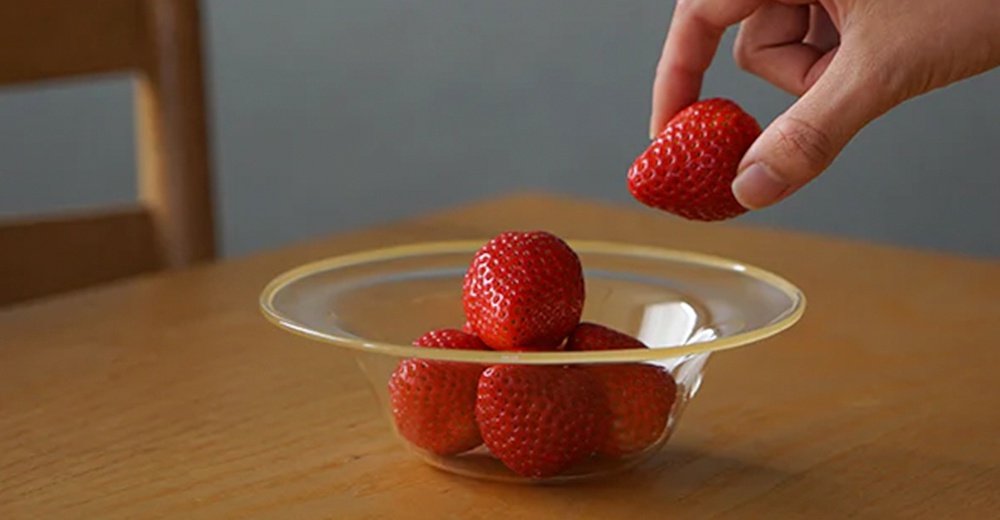 一個透明玻璃製成的深型點心碗，碗內盛有數顆新鮮的紅色草莓，一隻手正準備將一顆草莓放入碗中。