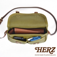 橄欖綠皮革傳統釣魚包，內部有深棕色皮革內袋，前袋放置手機及藍色圖案織品，搭配棕色皮革肩帶，包款為HERZ品牌。