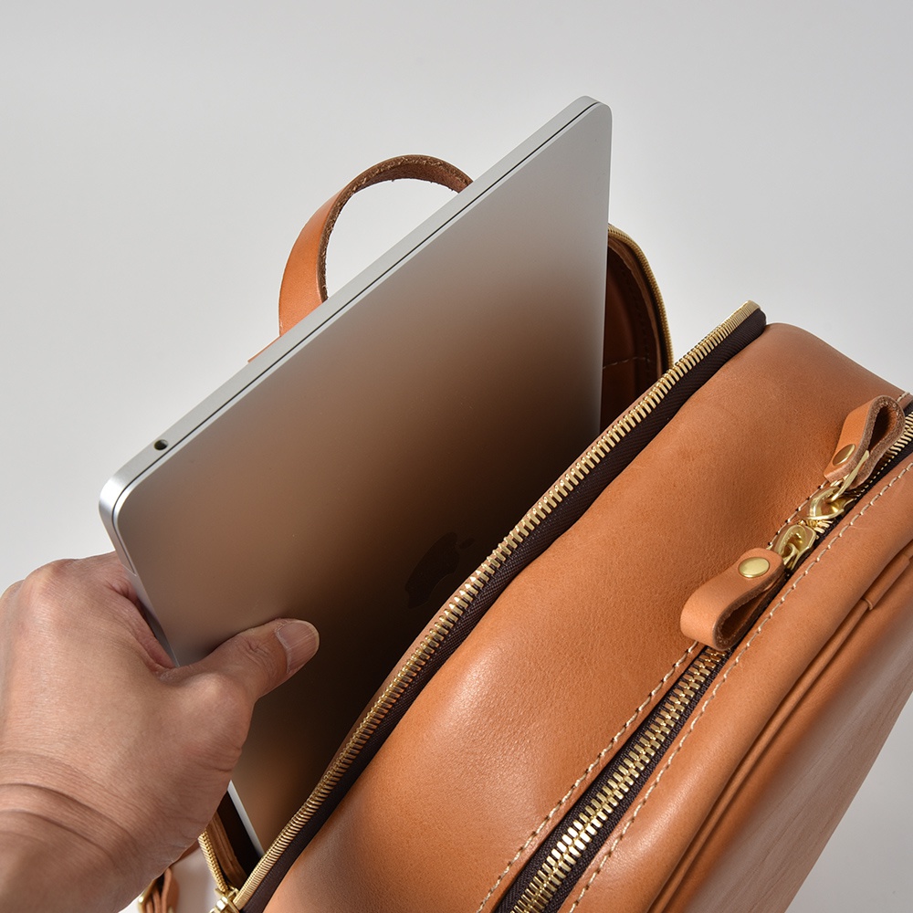 職人手作皮革多功能後背包 R-21，展示棕色皮革材質，一個銀色筆記型電腦正在被放入背包的拉鍊開口處。