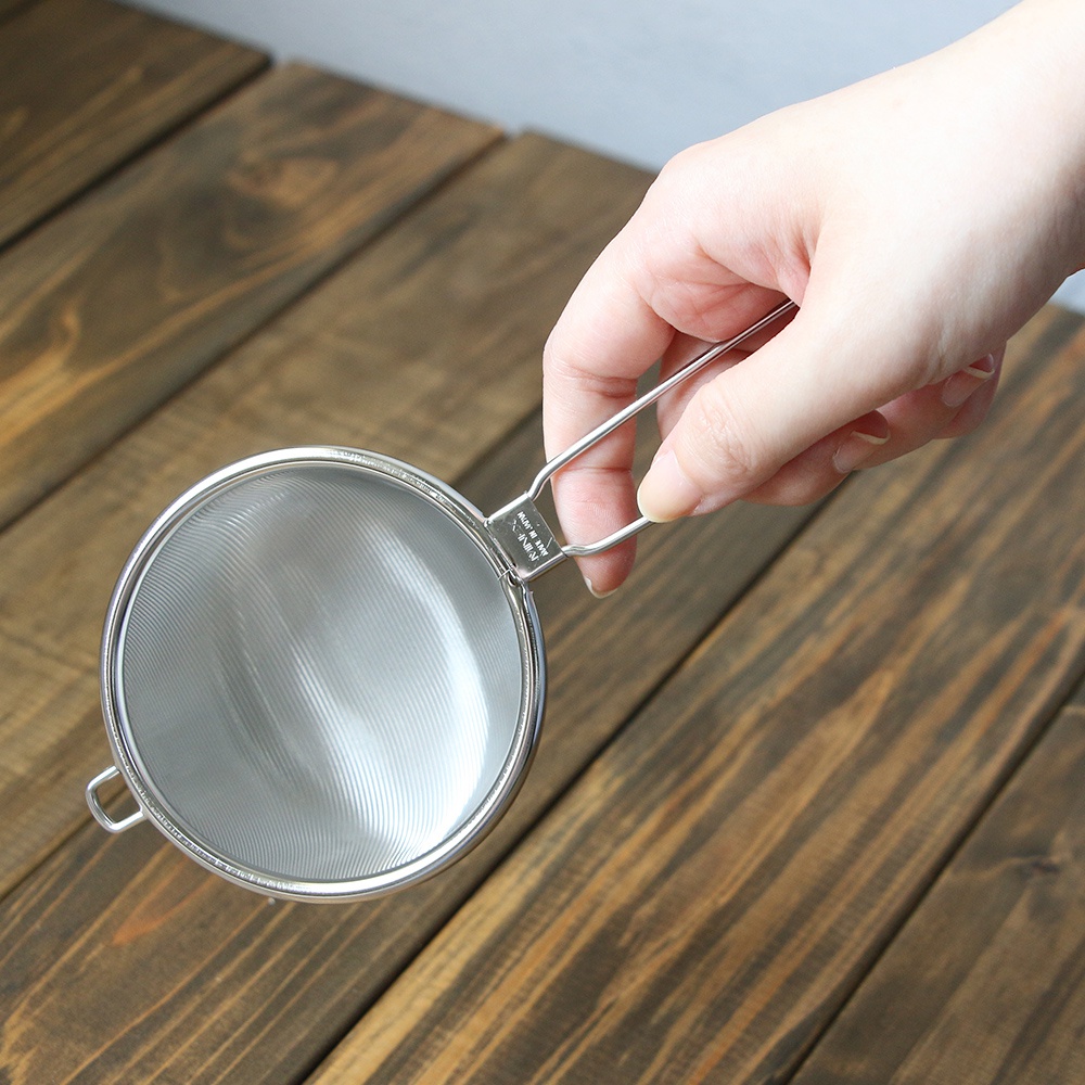 手持一個圓形、細網狀的不鏽鋼濾茶器，濾網為銀色，帶有細密的紋路，濾茶器外緣和手柄亦為不鏽鋼材質，手柄呈細長條狀，背景為深棕色木紋桌面。