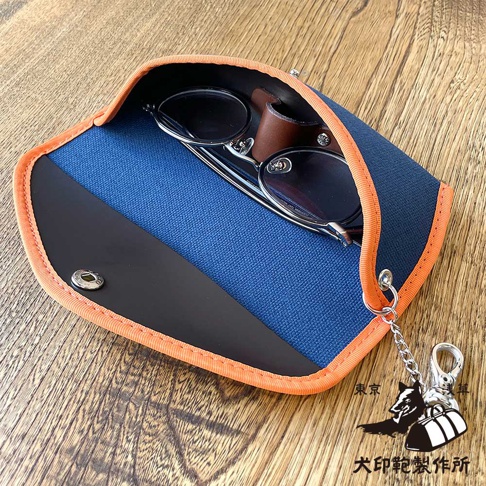 藍色帆布眼鏡收納盒，邊緣為橘色滾邊，內有眼鏡，掛繩上飾有犬印鞄製作所的標誌和日文文字。