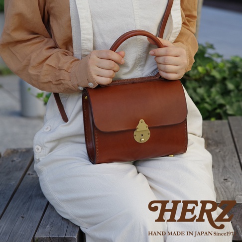 手提包為棕褐色皮革製成，配有金色金屬鎖扣和圓形提把，包身縫線明顯，風格復古。