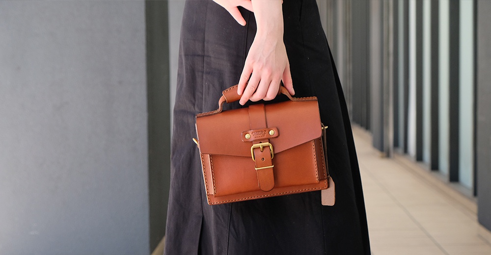 棕色皮革迷你單肩包，帶有翻蓋、金屬釦環和手提把，適合搭配黑色裙子。
