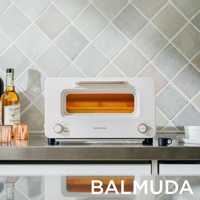 The white Balmuda The Toaster Pro K11C-SE sits on a stainless steel counter, with two slices of bread inside glowing orange.