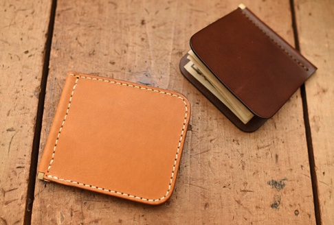 兩個職人手作皮革短夾，一個為淺棕色，一個為深棕色，皆為橫式對折設計，邊緣有白色車縫線，深棕色短夾內夾有鈔票。